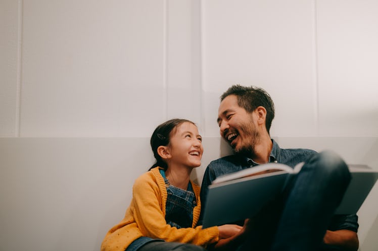 A dad tells his daughter a dad joke, laughing while reading a book to her.