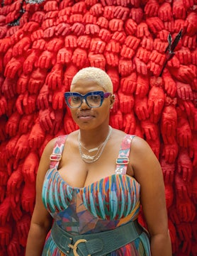 the artist Ebony G. Patterson in front of a sculpture made of red gloved hands titled "...fester..."...