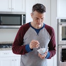 A man standing in his kitchen, eating a yogurt cup with a spoon.