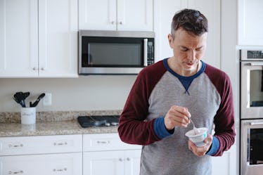 A man standing in his kitchen, eating a yogurt cup with a spoon.