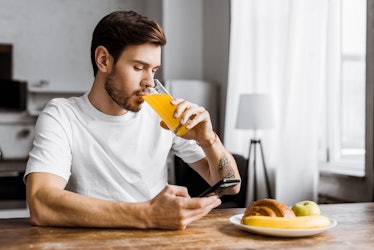 A man drinking apple juice while looking at his smartphone and eating breakfast.