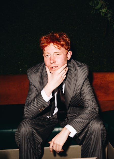 Archy sitting, holding his face, wearing a grey Etro suit