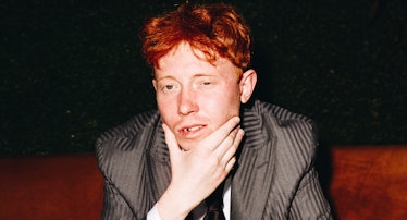 Archy sitting, holding his face, wearing a grey Etro suit