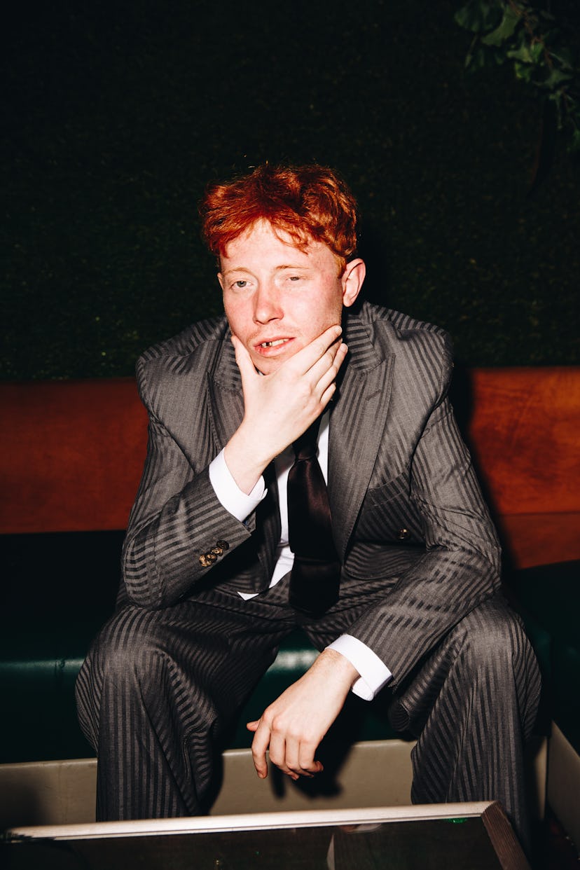 Archy sitting, holding his face, wearing a grey Etro suit