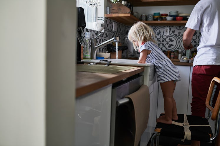 A girl standing on a chair and washing dishes in the kitchen sink.