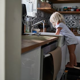 A girl standing on a chair and washing dishes in the kitchen sink.
