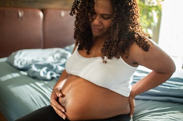 A pregnant woman with pregnancy farts sits on her bed and holds her lower back.