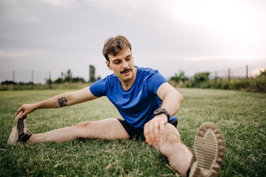 A man with gym dick working out, stretching his legs to the side while sitting in the grass.