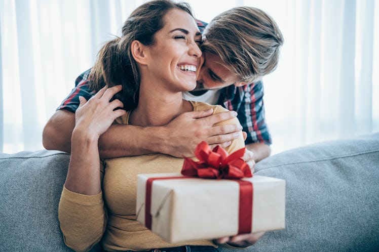 Woman with present on her lap sitting on a couch being hugged by a man,
