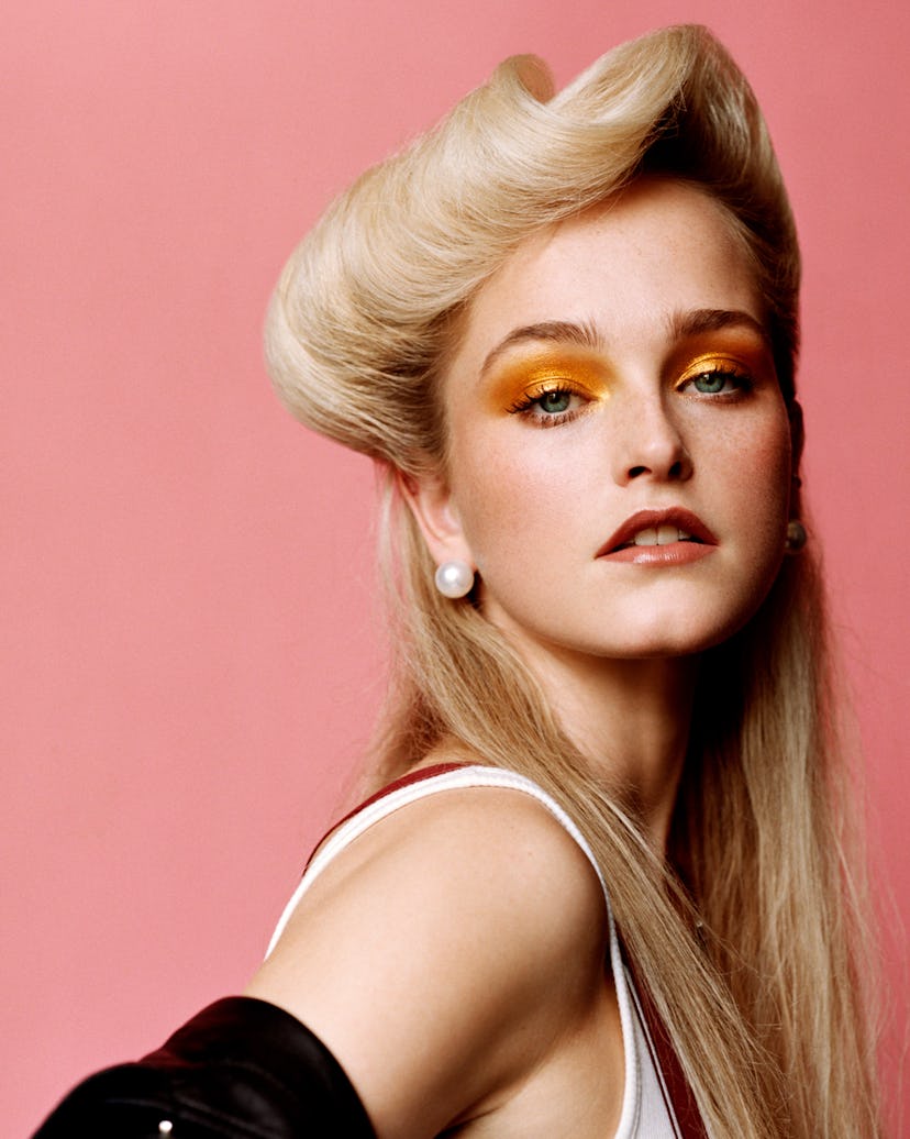 a blonde woman in front of a pink background with artfully swooped hairstyle and orange eyeshadow