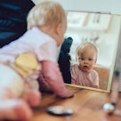 A baby crawling on the floor, looking in the mirror.