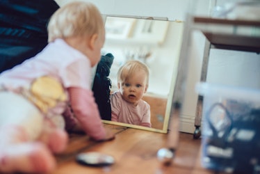 A baby crawling on the floor, looking in the mirror.