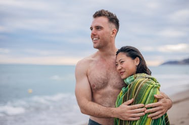 A hairy man with chest hair holding his girlfriend who is wrapped in a towel while they stand on a b...