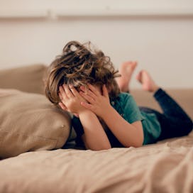 A child lying on a couch, covering their face with their hands.