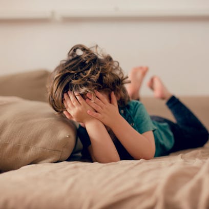 A child lying on a couch, covering their face with their hands.