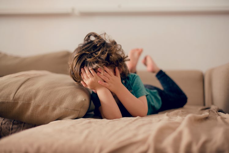 A child lying on a couch, covering their face with their hands.