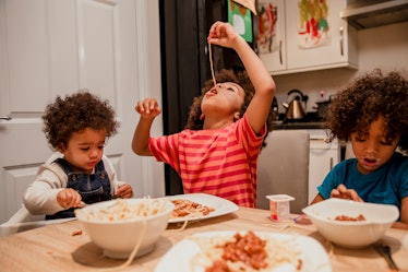 Three children at a dinner table eating spaghetti. One dangles a noodle into their mouth.