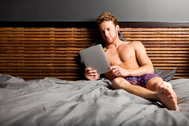 A man in his underwear on his bed, looking at his tablet.