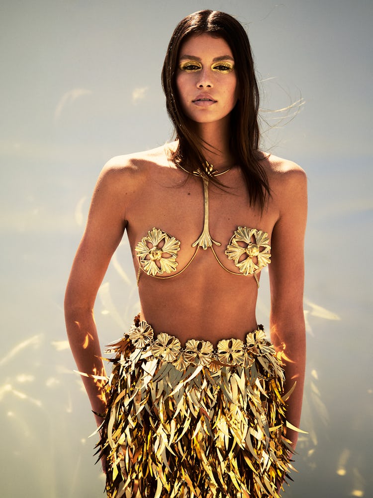 Model Kaia Gerber wears a gold bra top, gold sparkle skirt, and belt. 