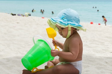 An August baby sitting on a beach, playing with a bucket and shovel.