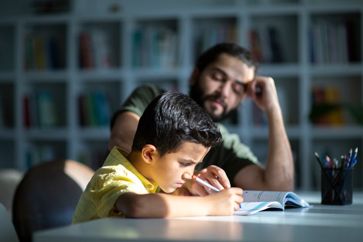 A dad leaning on his elbow helps his high-achieving son who is writing, working on a school assignme...