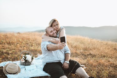 A fat man on a picnic with a woman outdoors in nature, taking a selfie.