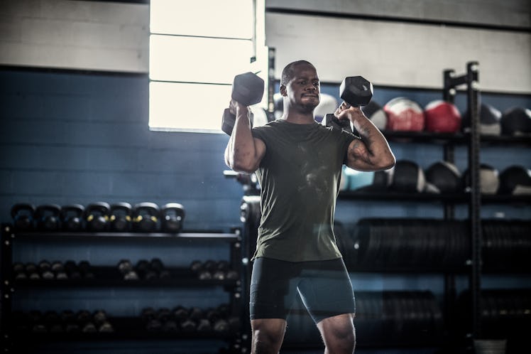 A man lifting dumbbells, doing a standing shoulder press, at the gym for strength training.
