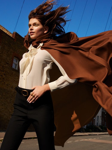 Model Kaia Gerber wears a brown cape, white blouse, black pants, belt, and rings.