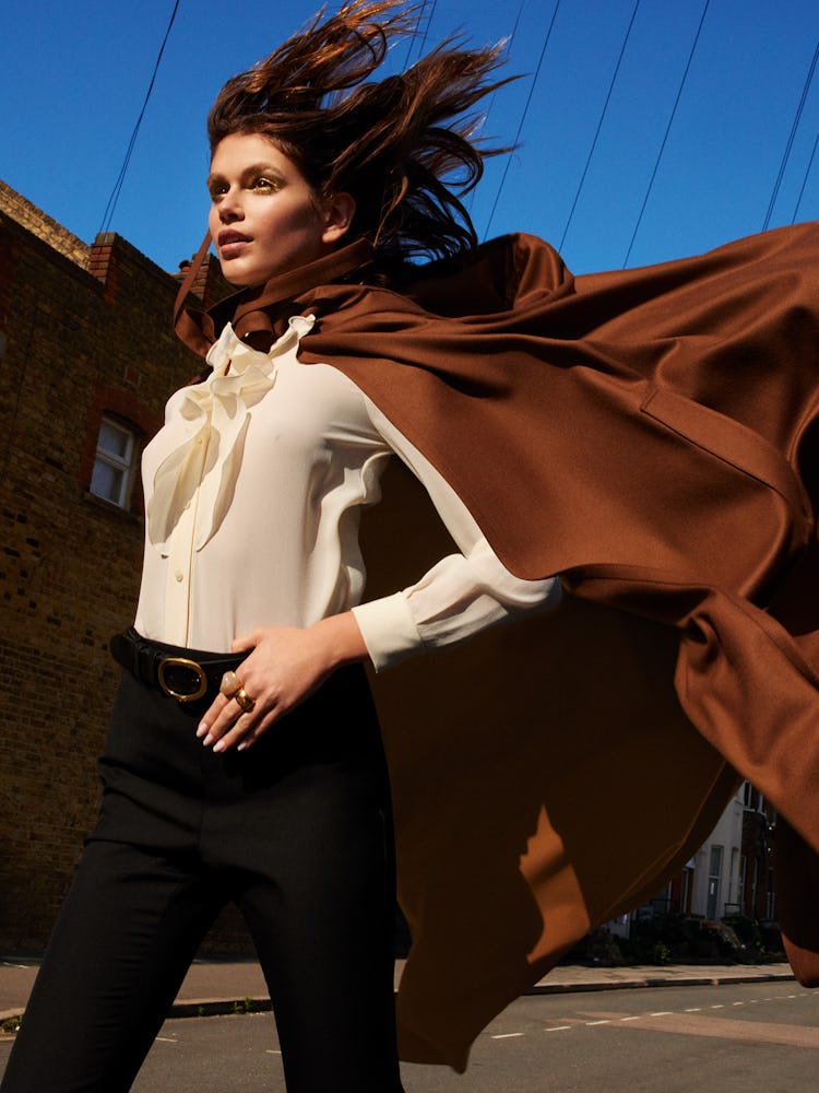 Model Kaia Gerber wears a brown cape, white blouse, black pants, belt, and rings.