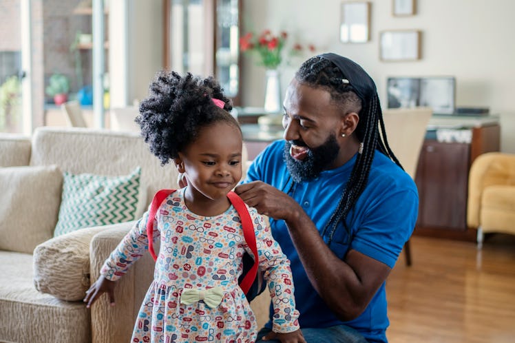 A dad helps his daughter get ready for Kindergarten by putting on her backpack at home.