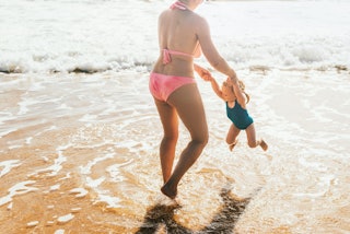 A woman in a bathing suit spins her child at the beach.