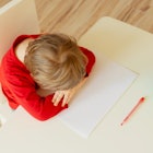 A Kindergartener in a red shirt putting their head on their desk.