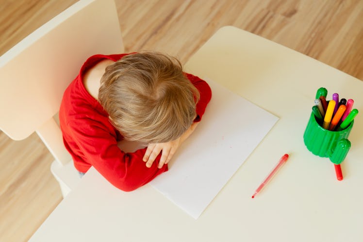 A Kindergartener in a red shirt putting their head on their desk.