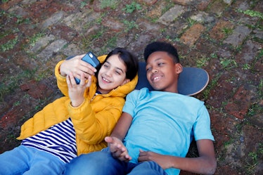 Two children lying on the ground with their heads resting on a skateboard, taking a selfie to post o...
