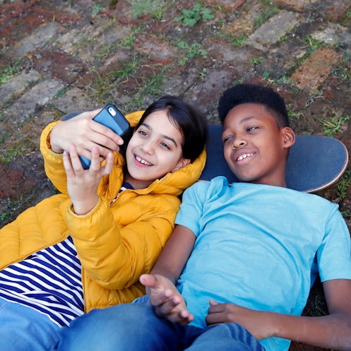 Two children lying on the ground with their heads resting on a skateboard, taking a selfie to post o...