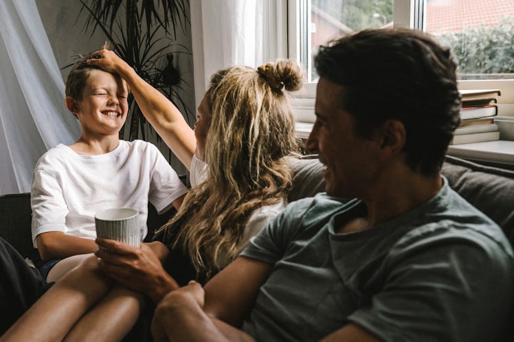 A mom and dad who are elephant parents ruffle their son's hair as they sit on a couch together.