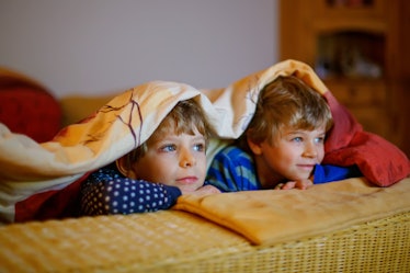 Two boys lying on a couch under blankets, watching TV.