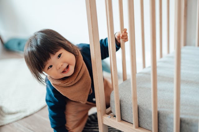 A smiling toddler peeks around the corner of their crib mattress.