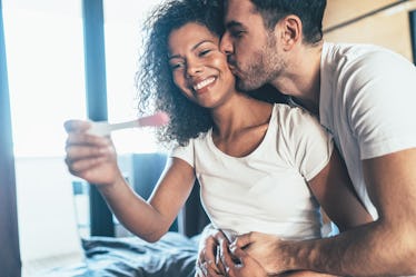 A man and woman holding a positive pregnancy test, looking happy as he kisses her cheek.