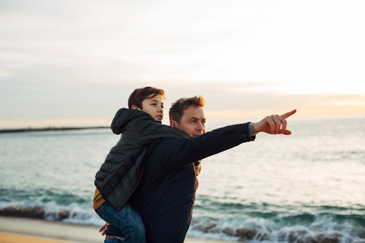 A dad giving his son a piggy back ride on the beach, in front of the sea, points off into the distan...