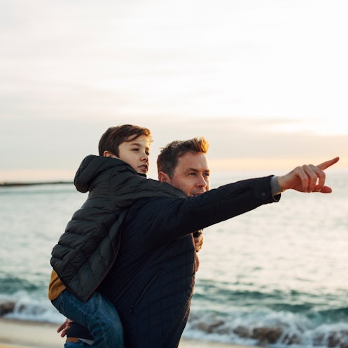 A dad giving his son a piggy back ride on the beach, in front of the sea, points off into the distan...