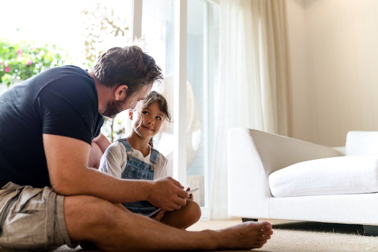 A dad and daughter sitting on the floor of their home, using a tablet.