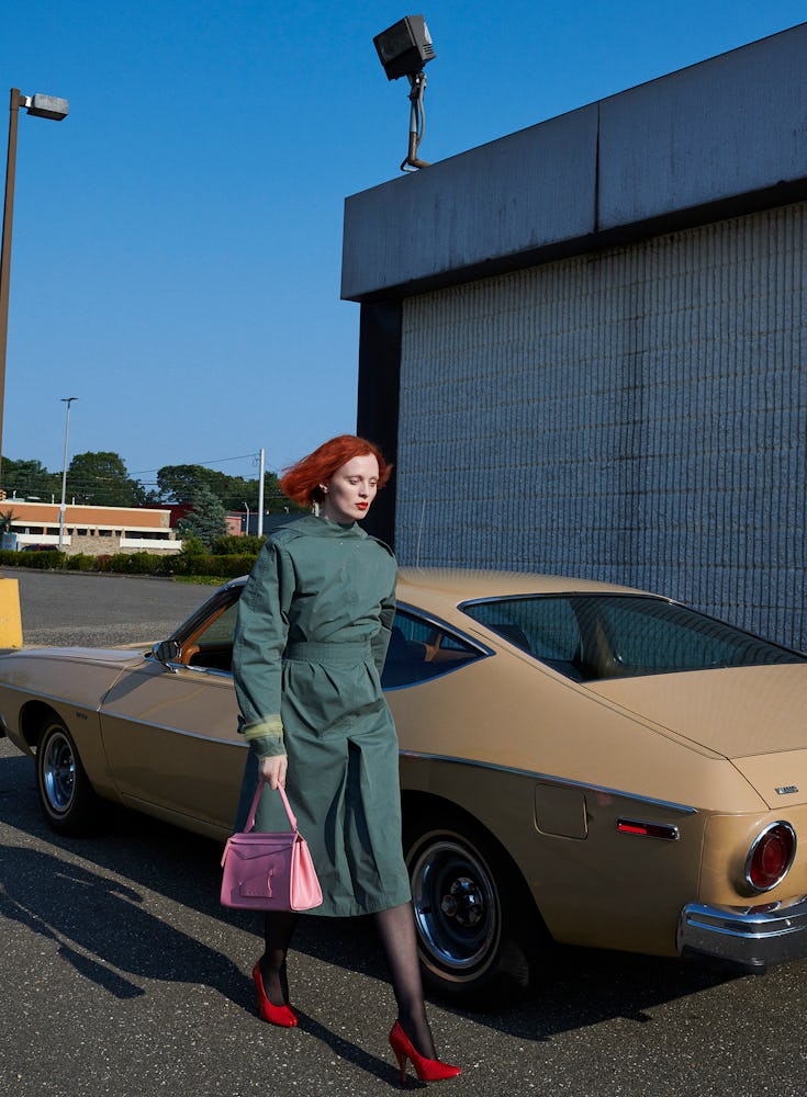 Model Karen Elson wears a dark green trenchcoat, pink leather bag, sheer socking, and red shoes.