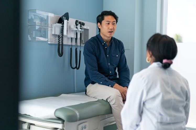 A man sitting on an examination table in a doctor's office talks to a medical provider in a white co...