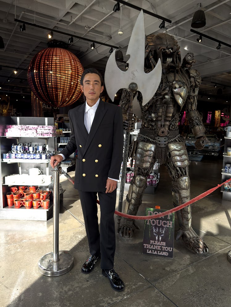Justin Min wears a black suit with gold buttons, white button down shirt and black shoes.