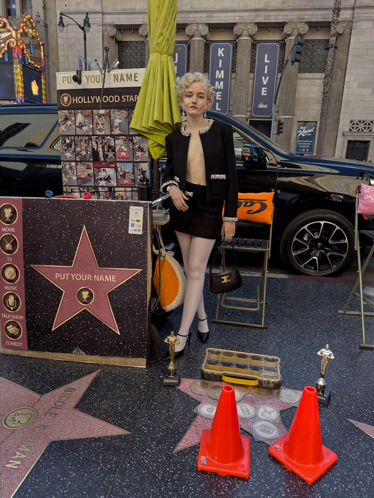 Julia Garner wears a Celine by Hedi Slimane jacket, skirt, bag, and shoes; Swarovski earrings; Carti...