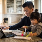 A dad works from home on his laptop at the kitchen counter while holding his toddler in his lap.