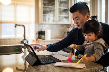 A dad works from home on his laptop at the kitchen counter while holding his toddler in his lap.
