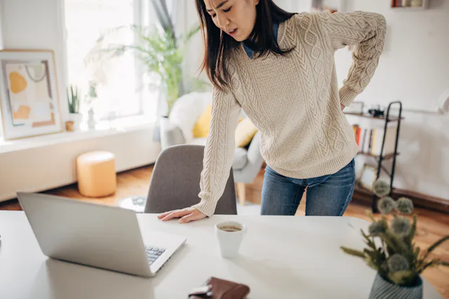 A woman clutches her lower back in pain as she gets up from her desk.