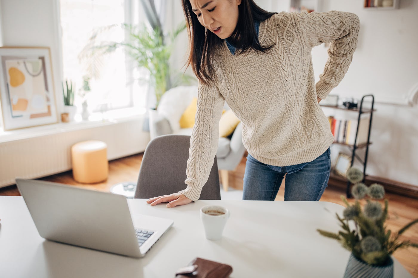 A woman clutches her lower back in pain as she gets up from her desk.
